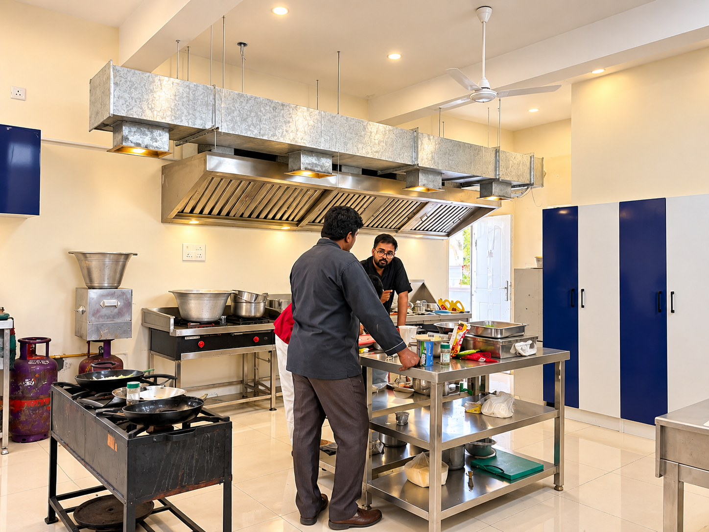 Chef guiding a trainee at the cooking range beneath the exhaust hood in the Fine Dine Academy kitchen